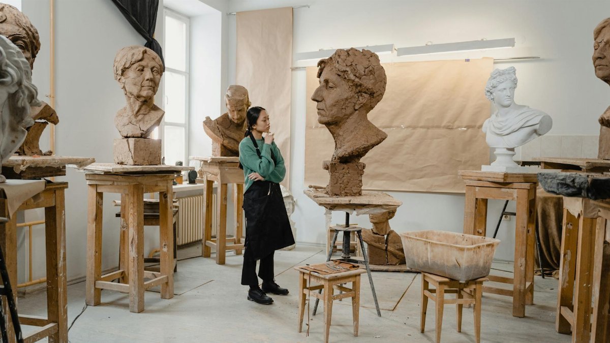 An artist in a creative clay sculpting studio surrounded by various sculpture busts on wooden stools.