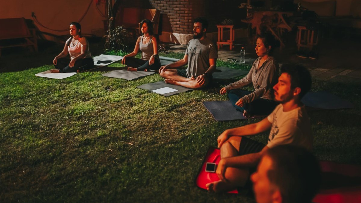 A diverse group meditating outdoors on yoga mats during evening time.