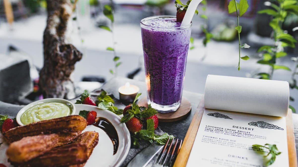 From above of glass of lavender creme brulee garnished with strawberry and plate of churro with avocado sauce placed on table with knife and fork and menu and candle against window decorated with plants