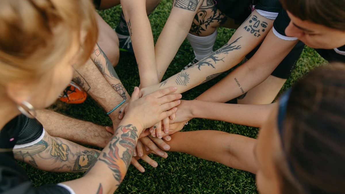 Tattooed team members join hands in a circle signifying unity and teamwork on a grassy field.