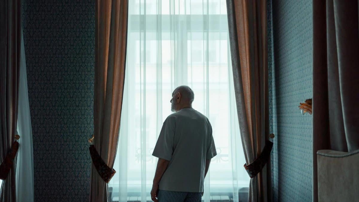 Elderly man standing by a window in an elegant room, looking outside. Ideal for concepts of isolation or contemplation.
