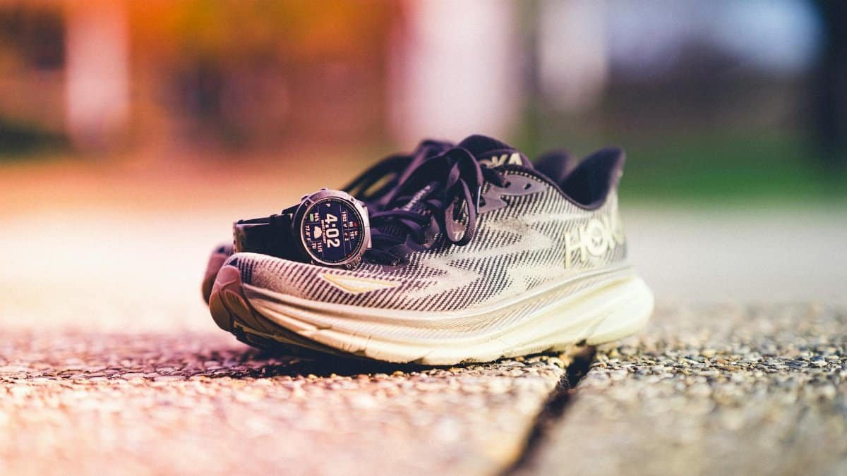 A pair of Hoka running shoes with a Garmin watch beside them on a sunny outdoor track.