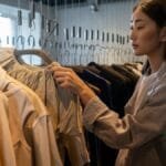 An Asian woman browsing clothing in a boutique shop, examining fashion items on hangers.