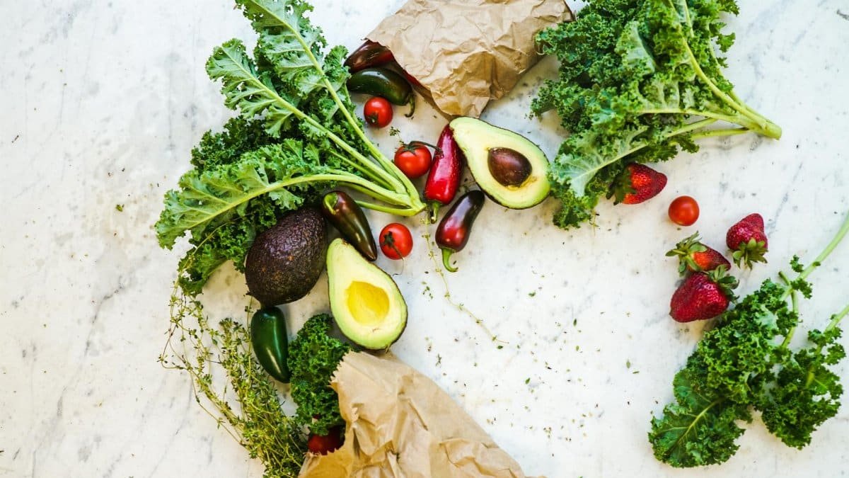Avocado, kale, and more fresh produce artfully arranged on a marble surface.
