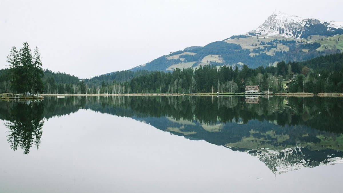 Picturesque landscape of calm lake reflecting rural house on bank and forest in front of mountain ridge covered with snow on cloudy day