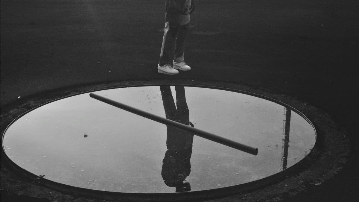 Monochrome image of a person reflected in a puddle wearing denim and sneakers outdoors.
