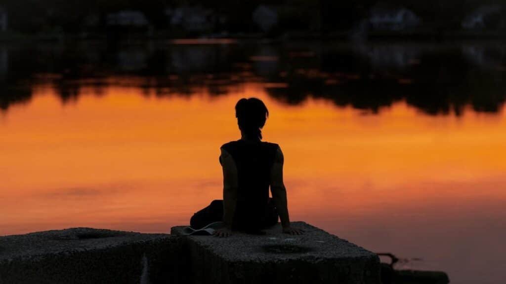 A person meditates at sunrise by a calm lake, creating a serene silhouette.