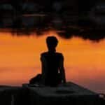 A person meditates at sunrise by a calm lake, creating a serene silhouette.