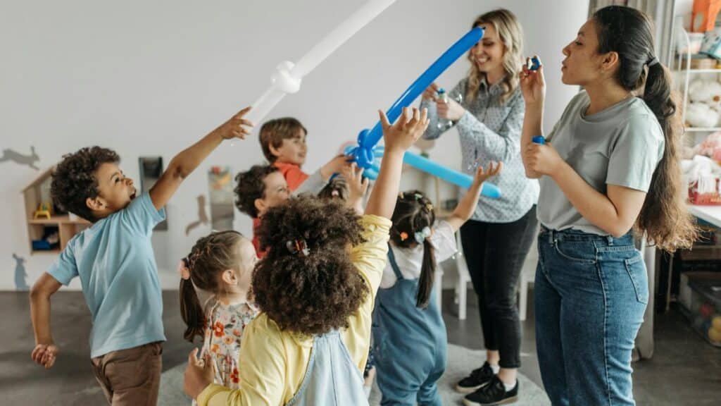 Group of children and teachers enjoying balloon games indoors in a kindergarten setting.