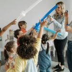 Group of children and teachers enjoying balloon games indoors in a kindergarten setting.