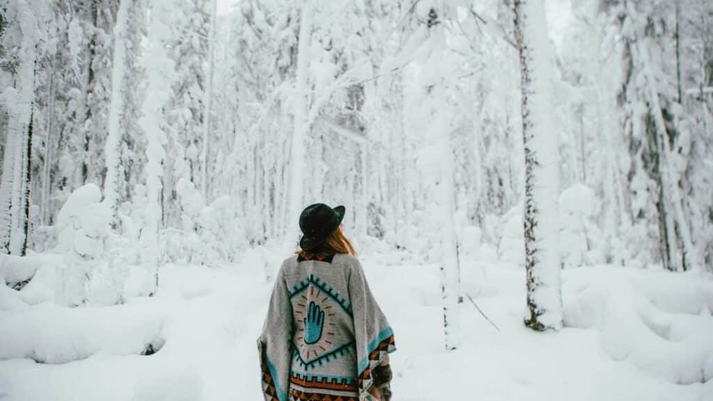 Back view of a woman in a poncho exploring a snow-covered forest during winter.