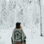 Back view of a woman in a poncho exploring a snow-covered forest during winter.