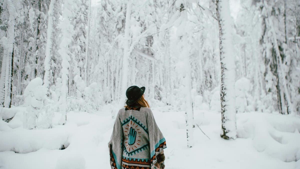 Back view of a woman in a poncho exploring a snow-covered forest during winter.