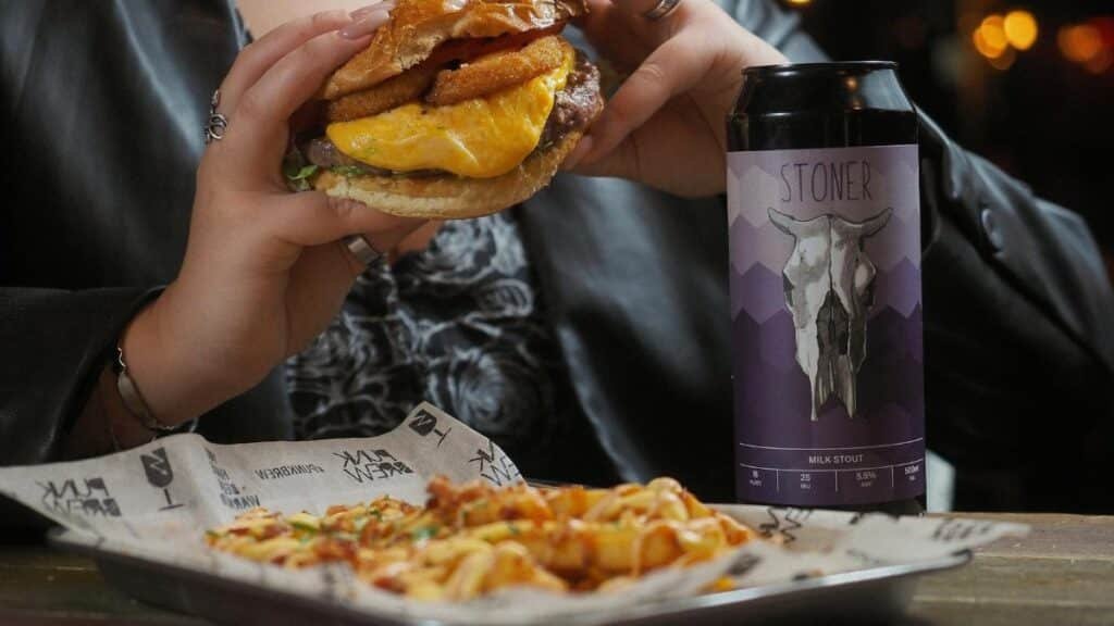 Close-up of a cheeseburger and beer can held by a person at a restaurant table.