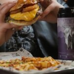 Close-up of a cheeseburger and beer can held by a person at a restaurant table.