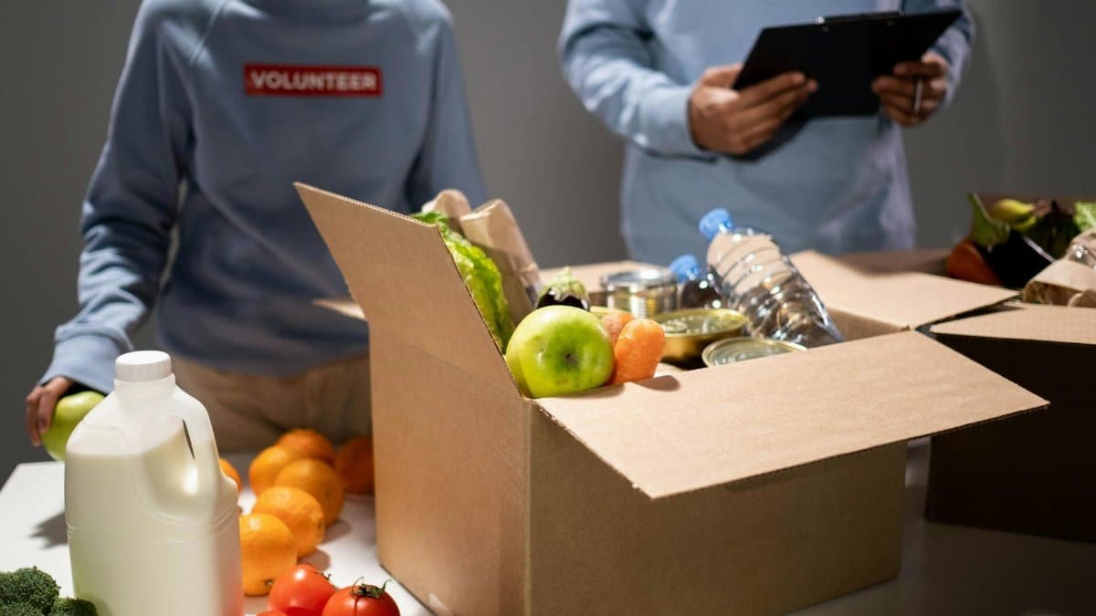 People packing a cardboard box with essentials like fruits, vegetables, and bottled water for charity.