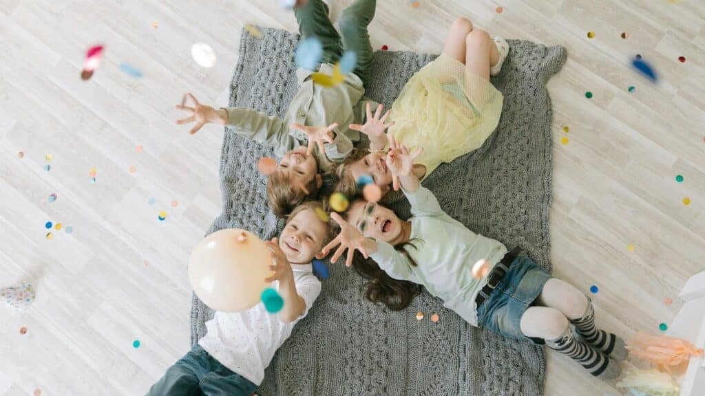 Aerial view of happy children celebrating with balloons and confetti indoors on a rug.