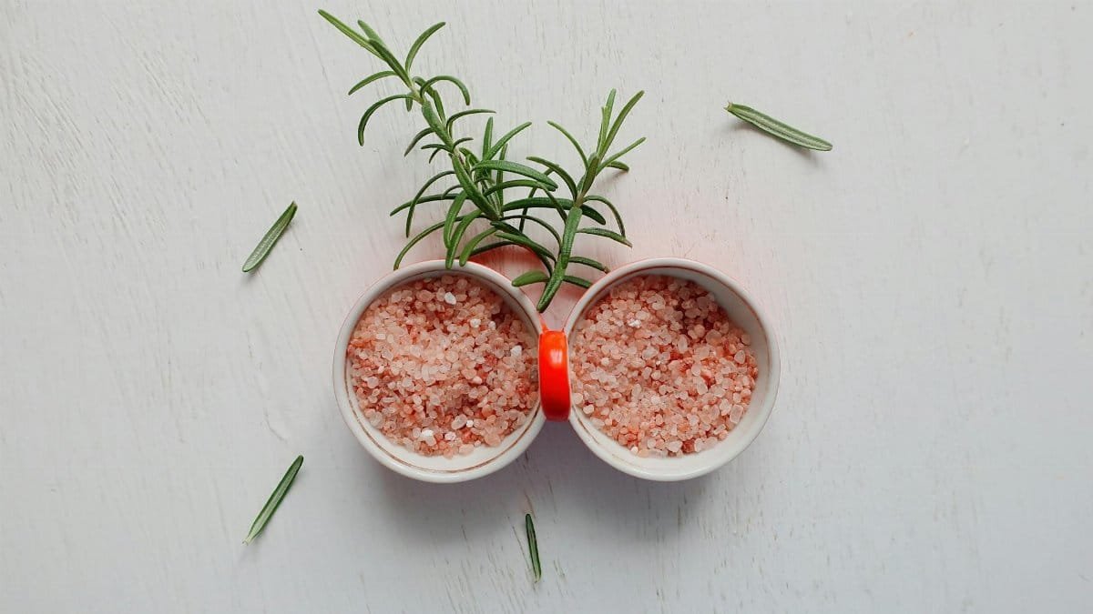 Top view of pink Himalayan salt and fresh rosemary in bowls on white table.