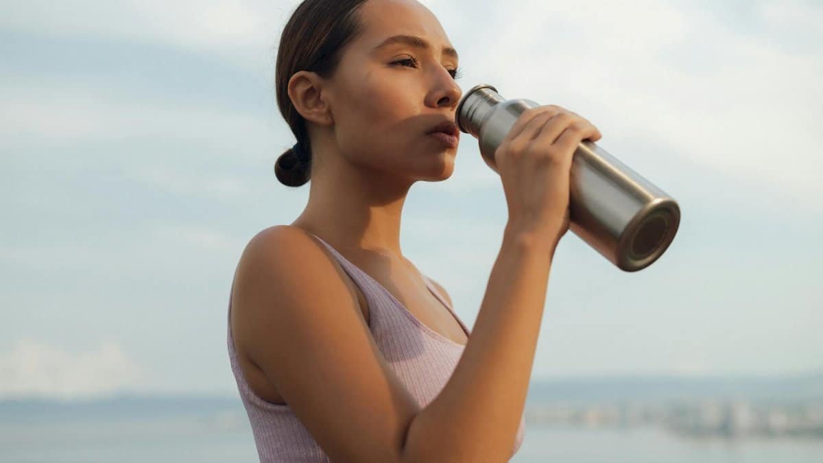 A woman drinks from a metal bottle on a sunny day by the beach, wearing a sports bra.