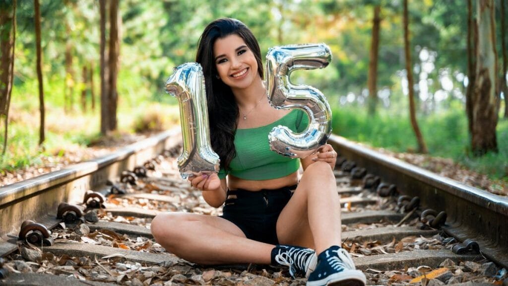 Teen girl sitting on railway tracks holding inflatable number 15 balloons, celebrating birthday outdoors.