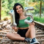 Teen girl sitting on railway tracks holding inflatable number 15 balloons, celebrating birthday outdoors.