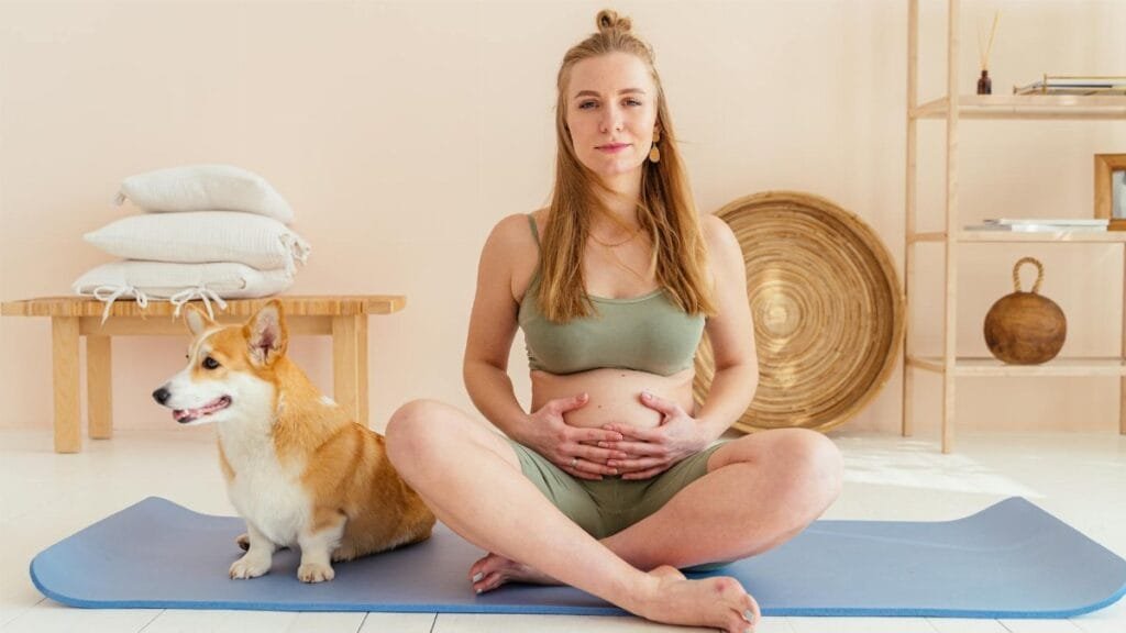 Pregnant woman sitting on yoga mat with a corgi in a cozy setting, embracing a healthy lifestyle.
