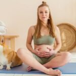 Pregnant woman sitting on yoga mat with a corgi in a cozy setting, embracing a healthy lifestyle.