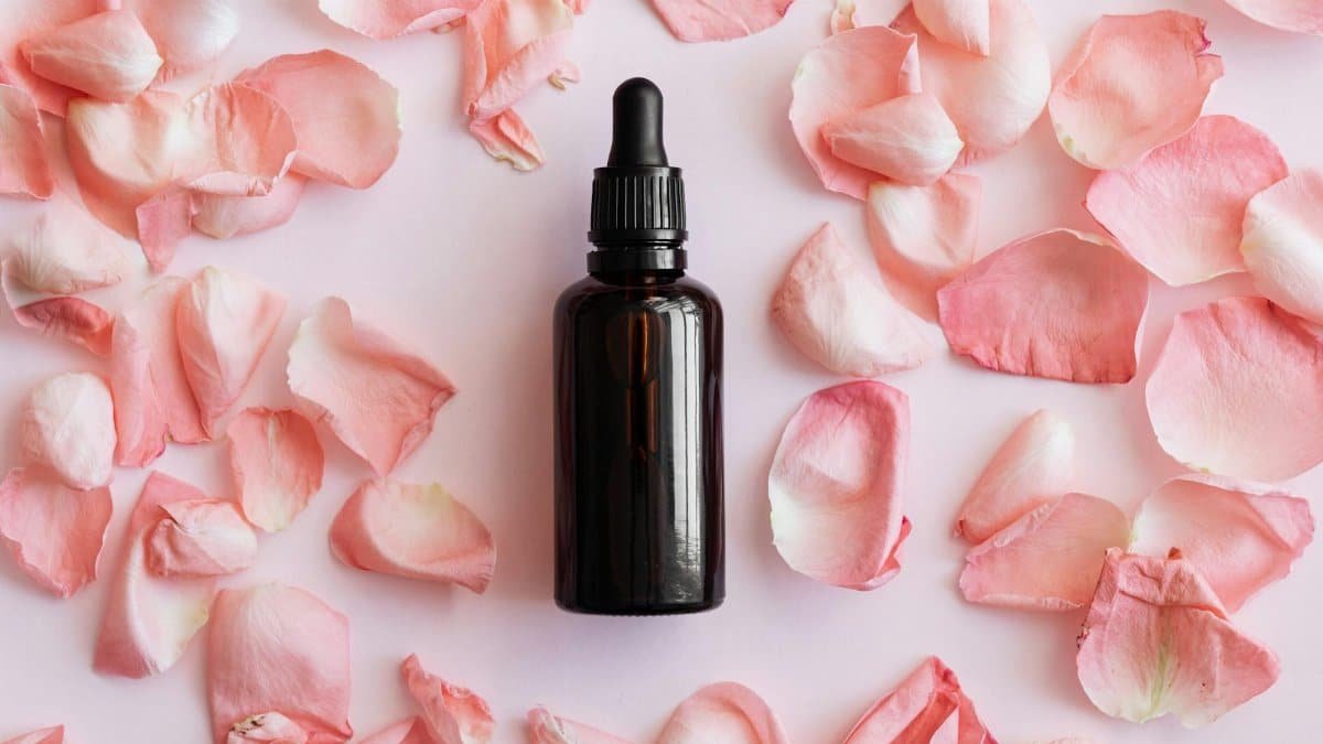 A dark glass dropper bottle surrounded by delicate pink rose petals on a pastel background.