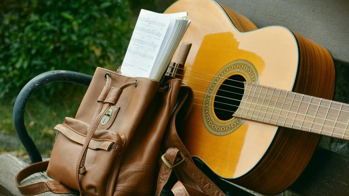 A serene outdoor scene of an acoustic guitar and a brown backpack on a wooden bench, perfect for music lovers.