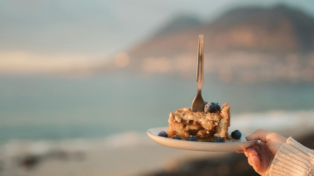 A scenic view of a blueberry pie slice with a fork, held by a hand at a beach.