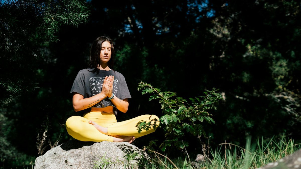 A woman practicing yoga outdoors, seated in a meditation pose on a rock, surrounded by lush greenery.
