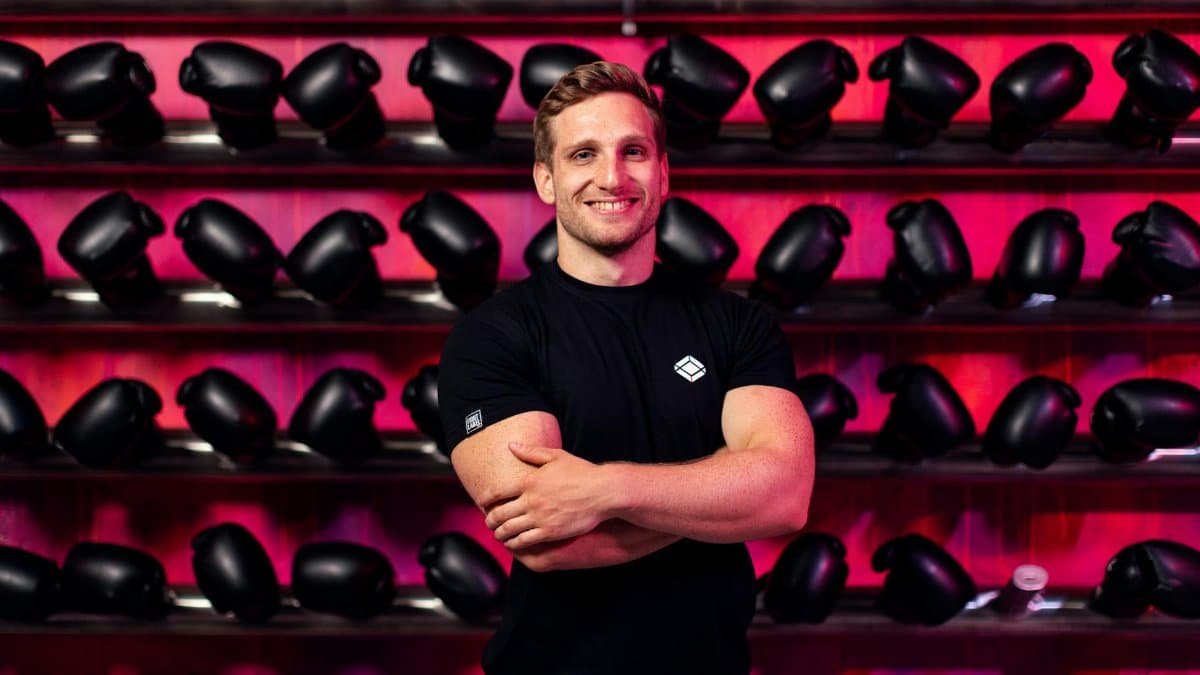 A smiling man in a gym stands confidently in front of a wall of boxing gloves.