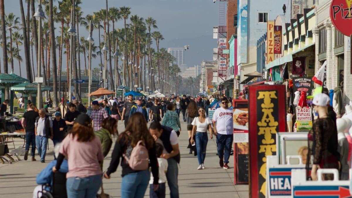 Crowds enjoying a sunny day on Venice Beach boardwalk lined with shops and palm trees.
