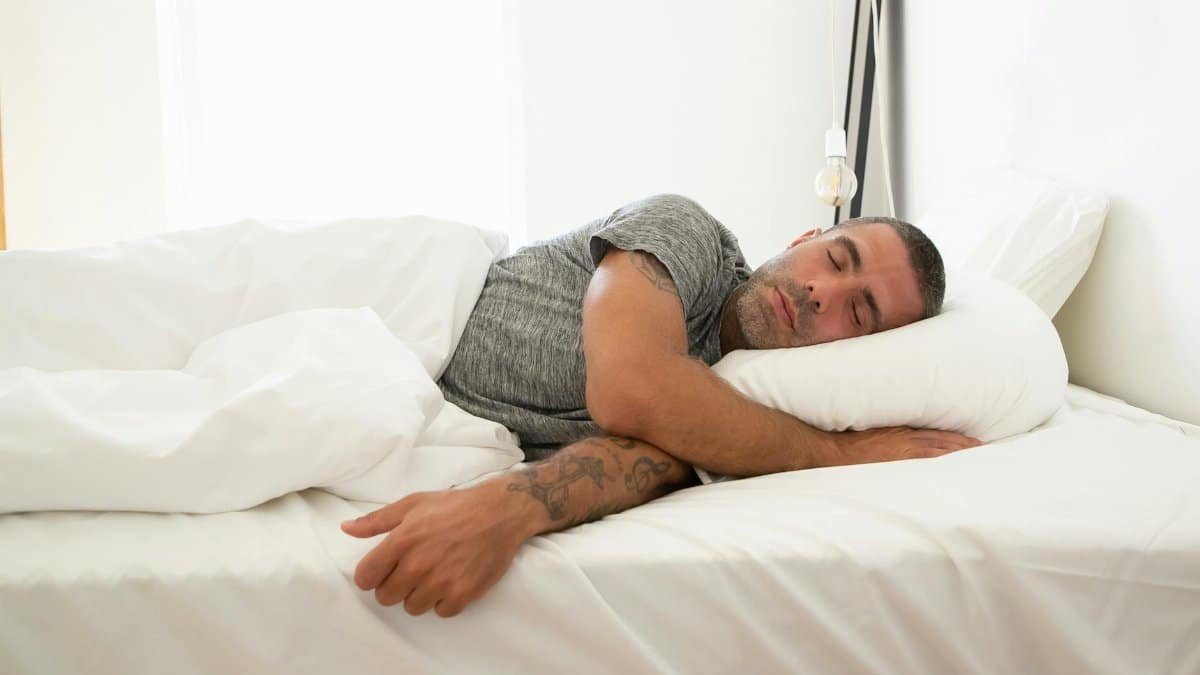 Adult man sleeping soundly on a white bed, tattoo visible, in a bright room.