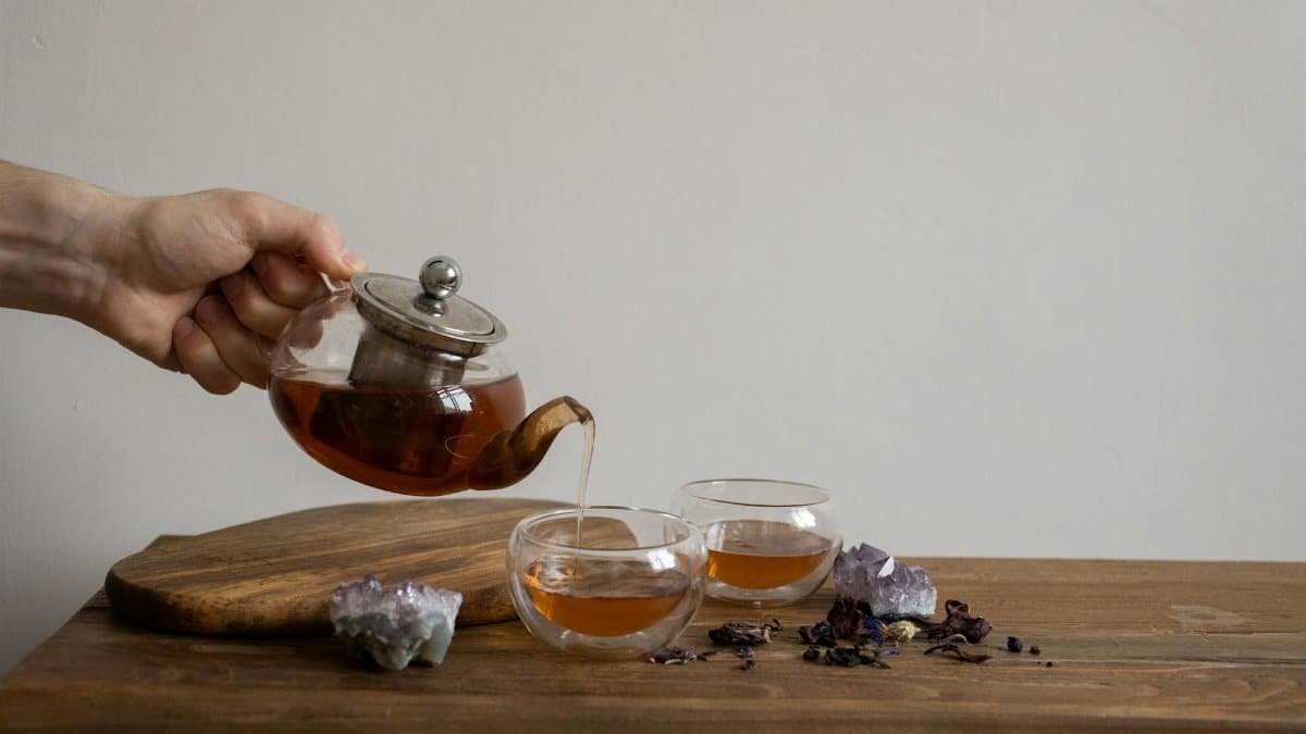 A hand pours tea from a kettle into glasses on a wooden tray with amethyst crystals.
