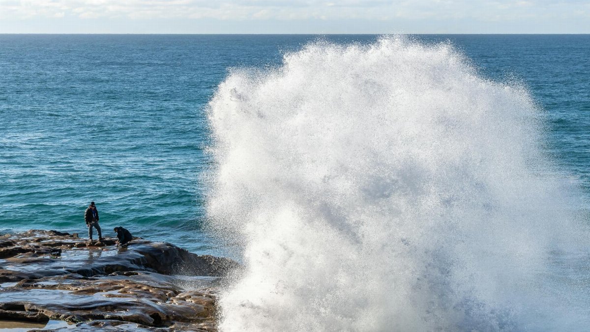 A powerful ocean wave crashes against rocky shore with people observing, capturing the dynamic scene.