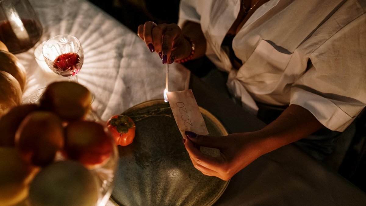 A woman sitting at a dining table, burning paper with a candle flame during a cozy evening ritual.