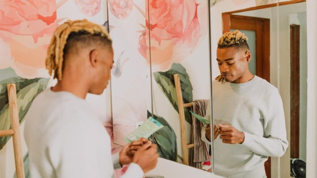 A man stands in a bathroom with floral decor, holding a skincare product and reflecting in the mirror.