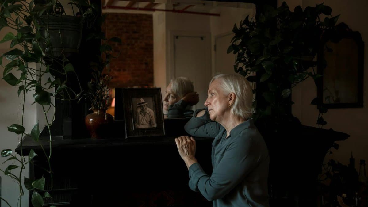 Elderly woman reflecting beside a photo frame and indoor plants.