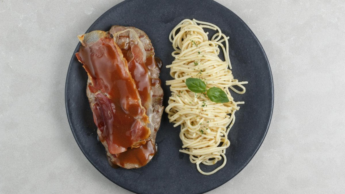 Top view of meatloaf with gravy and spaghetti garnished with basil on a black plate.