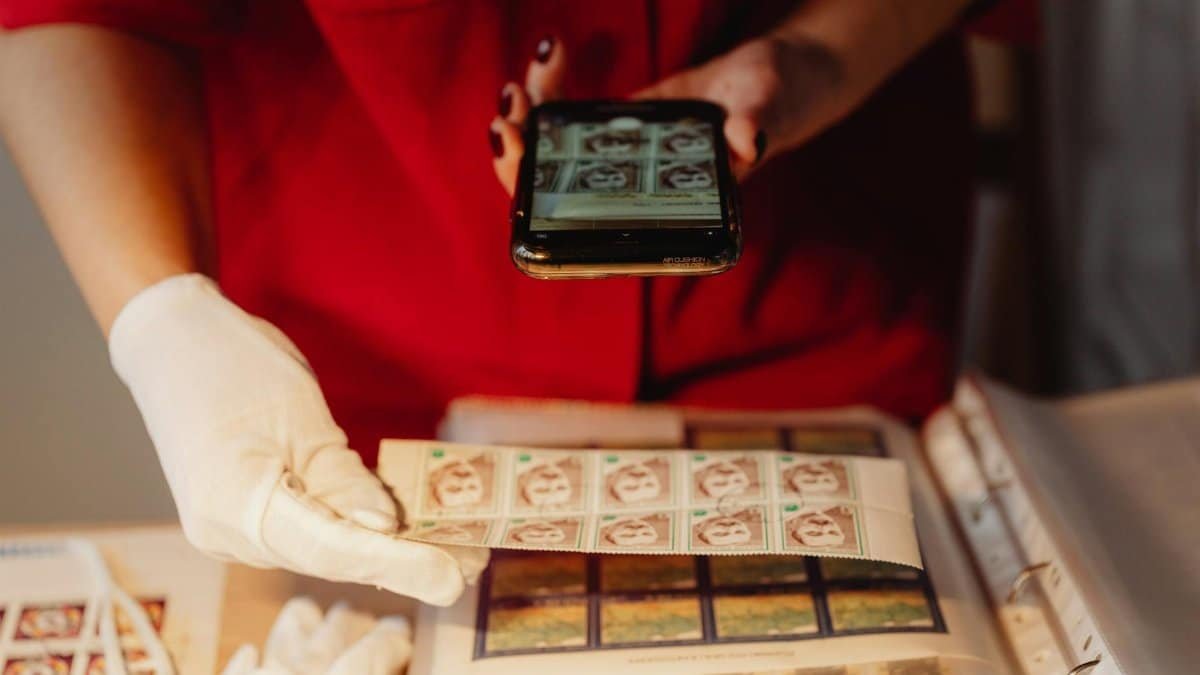 A person in gloves photographs a stamp collection using a smartphone for digital archiving.