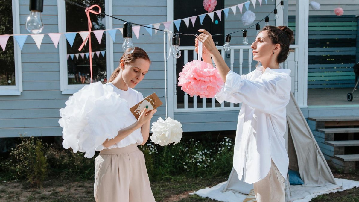 Two women in a backyard preparing party decorations with pom-poms and lightbulbs.
