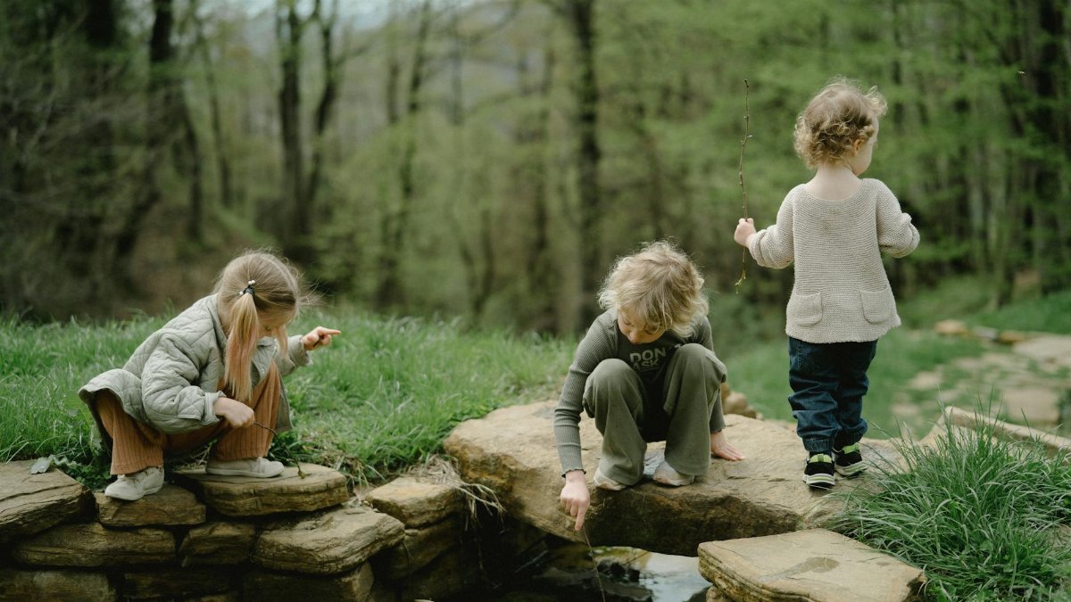 Three children playing by a stone-lined stream in a lush green forest during springtime.