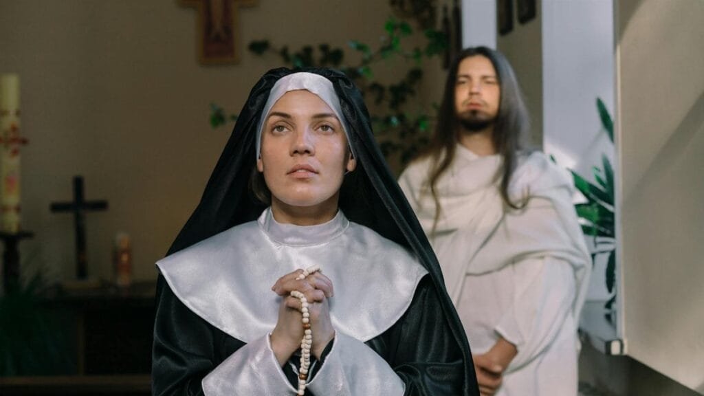A nun in prayer holding a rosary in a religious setting with a cross in the background.