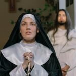 A nun in prayer holding a rosary in a religious setting with a cross in the background.