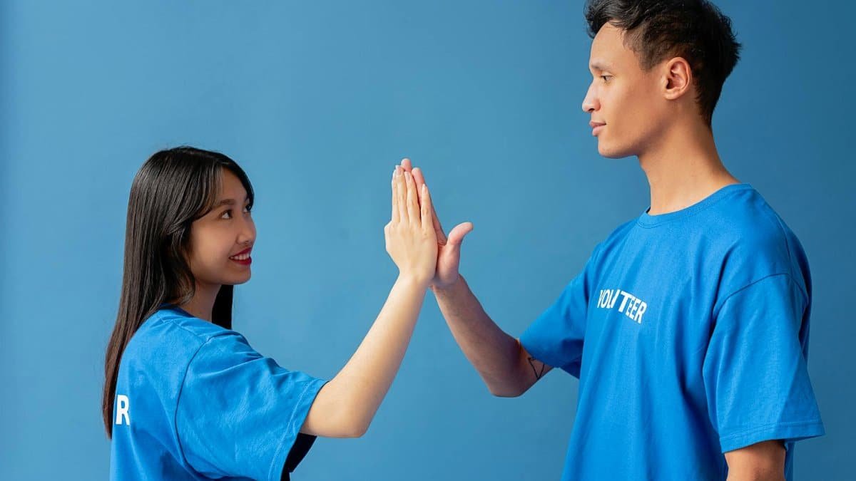 Two young volunteers in blue shirts high-five against a blue background, symbolizing teamwork.