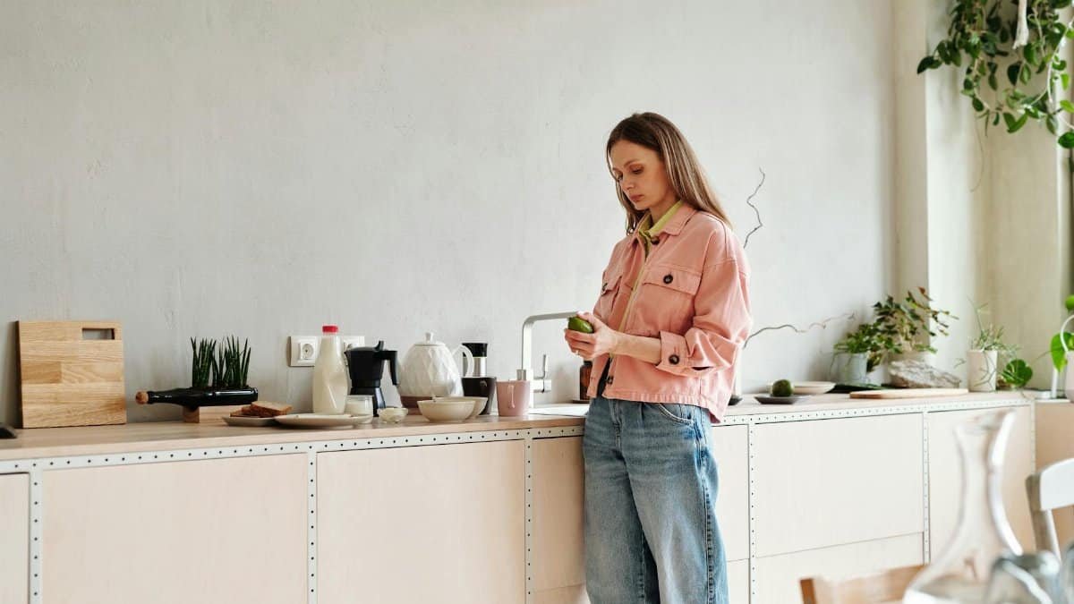 A woman stands in a modern kitchen with plants, holding an avocado.