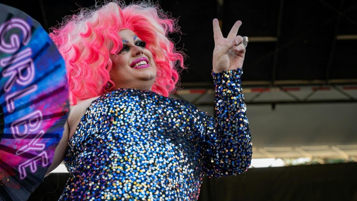 A glamorous drag queen with pink hair poses on stage holding a fan at a lively outdoor event.