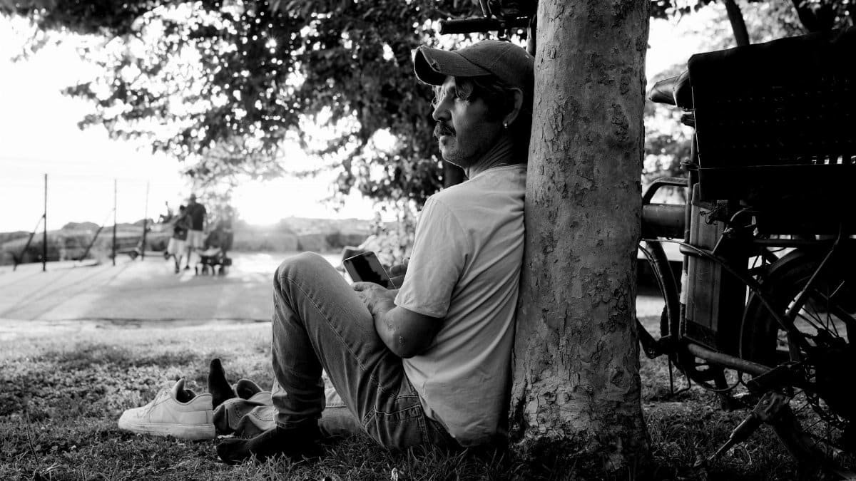 A man sits against a tree, lost in thought, in a tranquil black and white park setting.