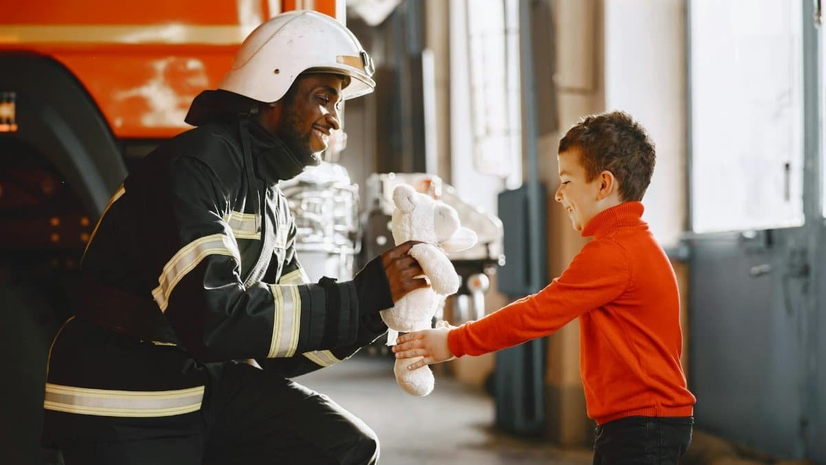 Firefighter in protective gear handing a teddy bear to a smiling young boy.